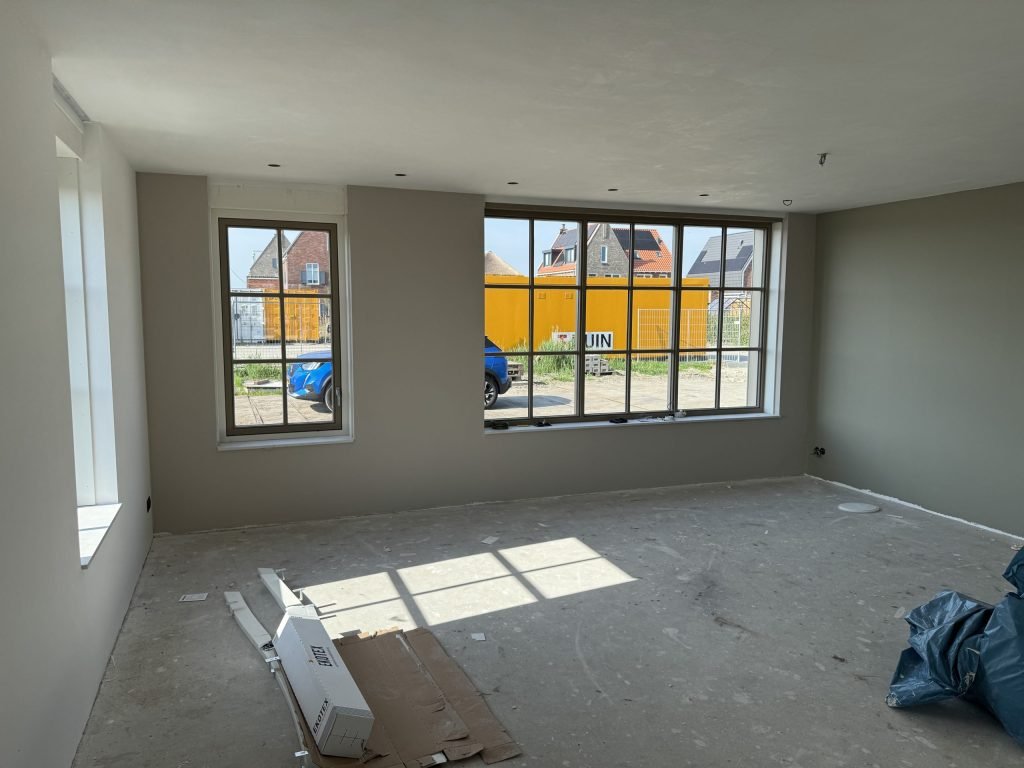 Lege nieuwbouwkamer met grijze wanden, een kale dekvloer en grote ramen met zicht op een bouwterrein