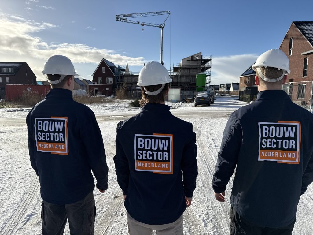 Drie vakmensen van Bouwsector Nederland met witte helm en bedrijfsjas, van achteren, voor een bouwkraan en huizen in aanbouw in de sneeuw